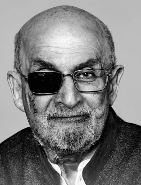 Head shot of a bearded Salman Rushdie after the knife attack. The right lens of his eyeglasses is blacked out and he has a knife scar on his right cheek. 