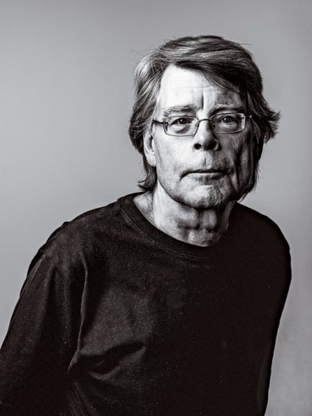 Black and white portrait of Stephen King from the waist up, with his hands behind his back, wearing an dark long-sleeved t-shirt and rectangular wire rimed vision correction glasses.