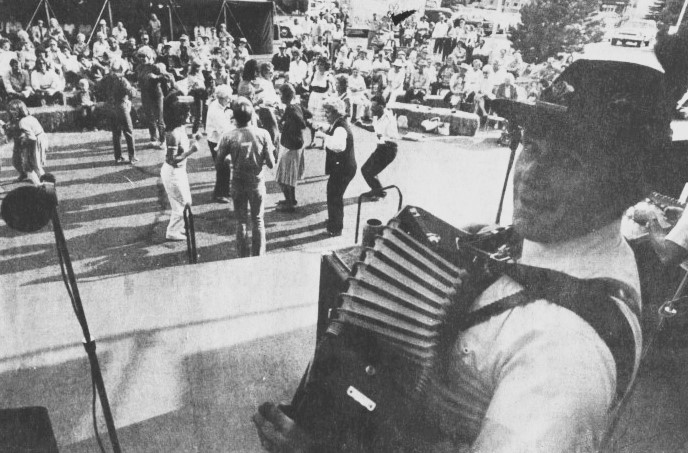 A man in traditional Bavarian dress plays the accordion while a group of people dance.