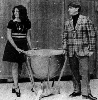 A woman and a man model fashionable outfits while leaning on a timpani.