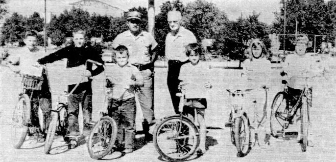 Six children pose with their bikes in front of a safety officer and another man.