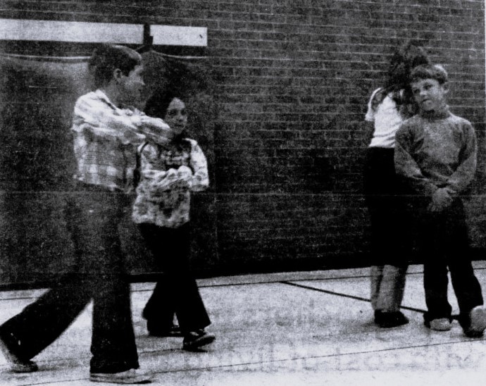 Four children square dance in the school gymnasium.