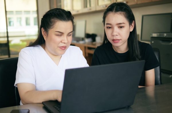 Two women sitting at a table using a laptop; one is visually impaired, and the other is assisting her.