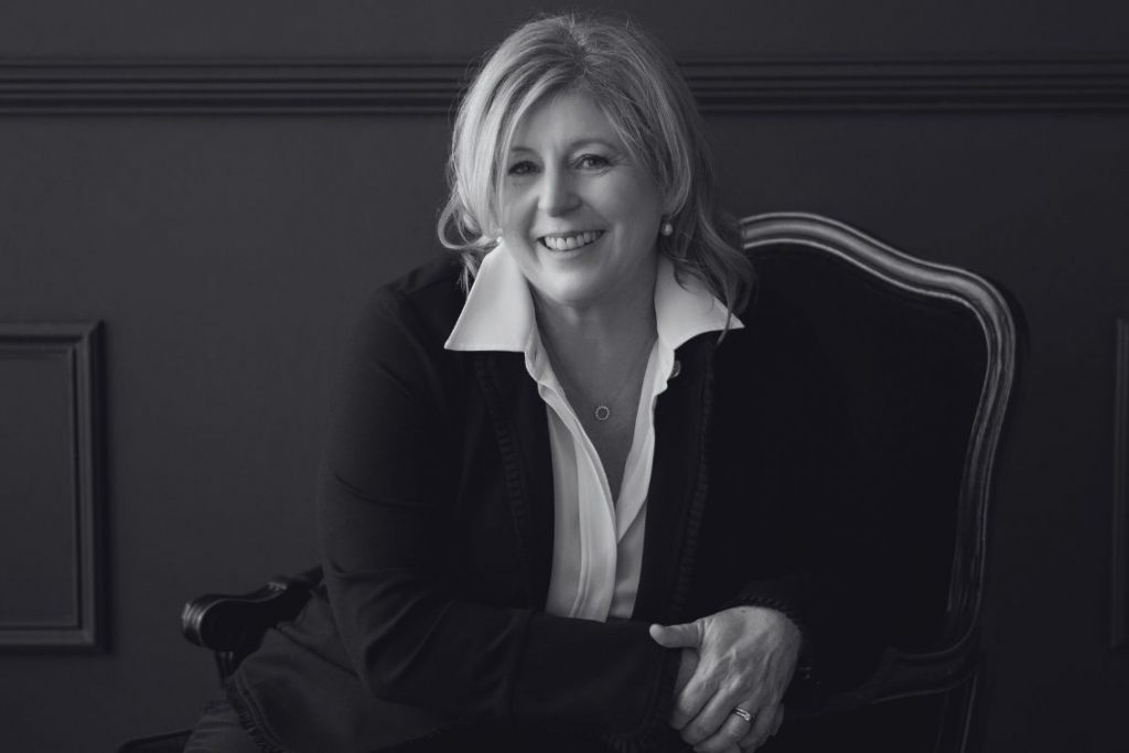 Black and white photo of Laine Moriarty sitting in a chair wearing a dark blazer and white button up shirt. Sitting in a chair directly facing the camera.
