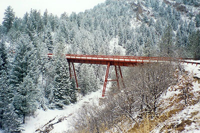 A red steel bridge spans across a snowy, wooded canyon. 