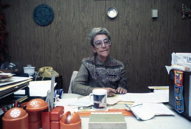 A librarian wearing a name tag sits at her office desk.