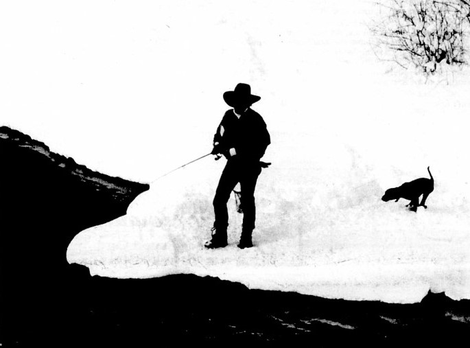 A man in a cowboy hat fishes on a snowy bank with his dog.