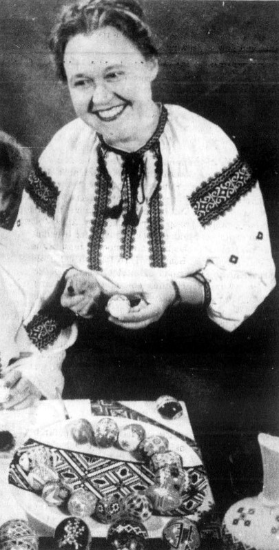 A woman in traditional Ukrainian clothing demonstrates an egg decorating technique.