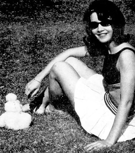 A young woman in sunglasses poses on the grass beside a tiny snowman.