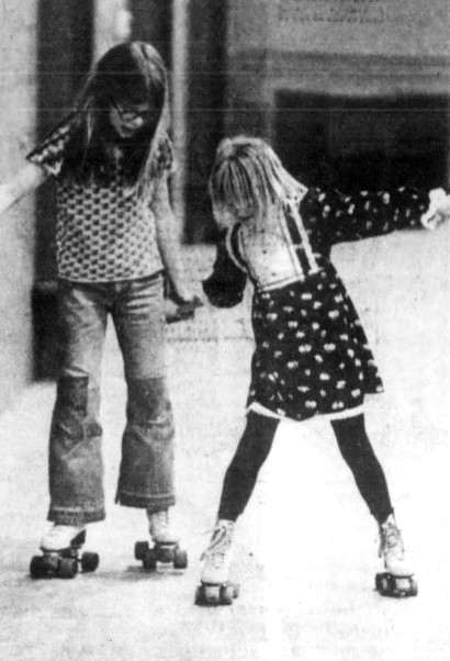 Two girls hold hands while they roller skate.