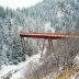 A red steel bridge spans across a snowy, wooded canyon.