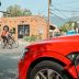 A red electric SUV plugged into an EV charger on a small town's main street while three people ride bikes in the background.